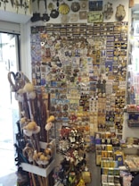 A shop display filled with an assortment of souvenirs, including Camino de Santiago badges, scallop shells, walking sticks, and various keychains. The wall is densely packed with colorful and detailed items, highlighting the theme of travel and pilgrimage.