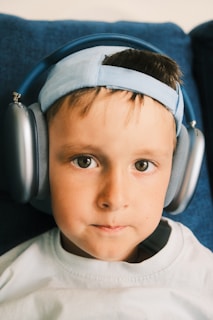 A child is wearing large over-ear headphones and a light blue baseball cap backwards. The child has brown eyes and is looking directly ahead. The background includes a dark blue cushion or sofa.