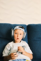 A young child sits on a blue couch, wearing large headphones and holding a video game controller. The child looks engaged and relaxed, with a light-colored t-shirt featuring a small palm tree design. The background is a light, textured wall.