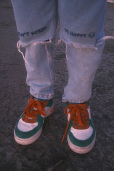 Worn and slightly torn light blue jeans with the phrases 'DON'T WORRY' and 'BE HAPPY' printed near the knees. The jeans are paired with colorful sneakers featuring green, orange, and white colors. The laces are bright orange, and the background appears to be a rough, textured ground surface.