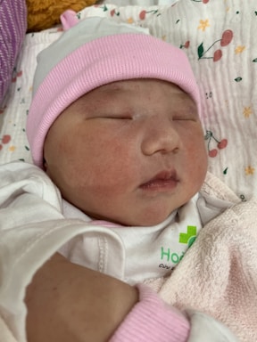 A newborn baby is peacefully sleeping, wearing a white and pink cap and wrapped in a soft blanket. The background contains a patterned fabric with small floral designs.