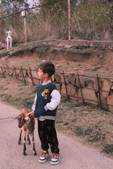 A young boy is standing on a paved pathway, holding a small goat on a leash. The boy is wearing a jacket with a letter 'E' and colorful patches, black pants with a pattern, and brown sneakers. There is a stone and wooden fence behind him, with a grassy area and another white goat grazing on a slope in the background. The setting appears to be outdoors with earth and greenery present.