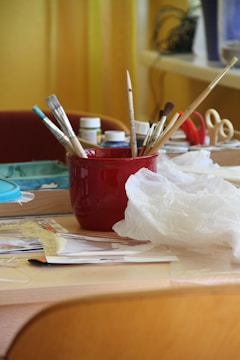 A red container holds various paintbrushes on a table cluttered with art supplies. Nearby, there are bottles of paint, a pair of scissors, and a plastic bag. The background features a window and some abstract shapes suggesting an indoor setting.