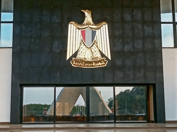 A large emblem depicting an eagle with outstretched wings, set against a dark wall. Below the emblem is a reflective glass surface, which shows the reflection of a monument with a triangular shape and greenery in the background.