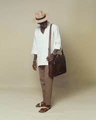 A stylishly dressed individual wearing a beige hat, white long-sleeved shirt, tan trousers, and brown sandals. They are holding a brown leather shoulder bag and have various accessories on their wrists.