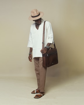 A stylishly dressed individual wearing a beige hat, white long-sleeved shirt, tan trousers, and brown sandals. They are holding a brown leather shoulder bag and have various accessories on their wrists.
