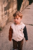 A young child with curly brown hair stands on a concrete path beside a textured, aged wall. The child is wearing a hooded jacket with multi-colored sleeves and brown corduroy pants, exuding a contemplative expression.