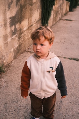 A young child with curly brown hair stands on a concrete path beside a textured, aged wall. The child is wearing a hooded jacket with multi-colored sleeves and brown corduroy pants, exuding a contemplative expression.