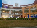 A shopping mall with a large facade, featuring prominent signage for HSBC and other stores. The building has multiple levels with visible shops. The area is landscaped with palm trees and trimmed shrubs. There are people walking in the foreground, suggesting a busy or bustling atmosphere.