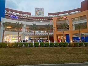 A shopping mall with a large facade, featuring prominent signage for HSBC and other stores. The building has multiple levels with visible shops. The area is landscaped with palm trees and trimmed shrubs. There are people walking in the foreground, suggesting a busy or bustling atmosphere.