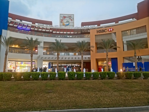A shopping mall with a large facade, featuring prominent signage for HSBC and other stores. The building has multiple levels with visible shops. The area is landscaped with palm trees and trimmed shrubs. There are people walking in the foreground, suggesting a busy or bustling atmosphere.