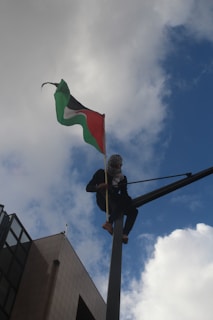 A person wearing a headscarf and dark clothing is perched high on a pole, holding a flag that is billowing against a backdrop of a blue sky with scattered clouds. The flag has green, black, white, and red colors. There is a building visible in the lower left corner of the image.