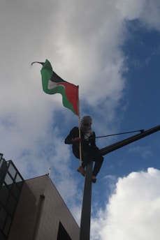 A person wearing a headscarf and dark clothing is perched high on a pole, holding a flag that is billowing against a backdrop of a blue sky with scattered clouds. The flag has green, black, white, and red colors. There is a building visible in the lower left corner of the image.