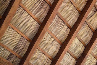 A close-up view of a thatched roof, composed of dried plant materials arranged in an orderly pattern between wooden beams. The materials have an aged appearance with varying shades of tan and brown, indicating a traditional, rustic architectural style.