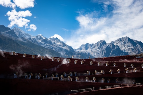 A group of people dressed in traditional clothing appear on multi-tiered platforms set against a backdrop of majestic snow-capped mountains under a bright blue sky with scattered clouds.