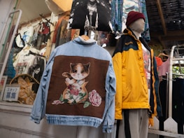 An assortment of clothes is displayed in a shop setting. A denim jacket with a large patch featuring a whimsical cat design is prominent. Beside it, a mannequin is dressed in a yellow and black jacket with a maroon beanie. Multiple shirts and decorative items hang in the background.