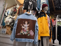 An assortment of clothes is displayed in a shop setting. A denim jacket with a large patch featuring a whimsical cat design is prominent. Beside it, a mannequin is dressed in a yellow and black jacket with a maroon beanie. Multiple shirts and decorative items hang in the background.