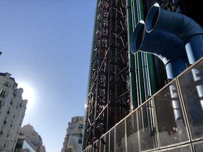 A large, industrial building with exposed colored pipes and a steel framework dominates the scene. The architecture features two prominent blue ducts or vents on the right side. On the left, a sunlit, old-style European residential building lines the street.