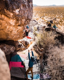 A climber is scaling a large boulder in a rocky outdoor setting, with a vast arid landscape in the background. Several people are observing and spotting the climber, standing on crash pads below. The scene captures a sense of adventure and outdoor activity.