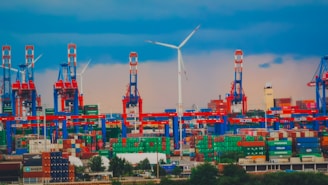 A busy port with numerous shipping containers stacked in various colors, large cranes towering over them, and a prominent wind turbine in the background against a cloudy sky.