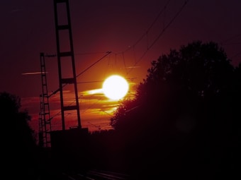 A vibrant sunset or sunrise with the sun glowing brightly against a colorful sky. Tall structures resembling power lines or railway poles stretch vertically, silhouetted against the sun. Trees frame the sun on either side, adding a natural element to the urban outline.