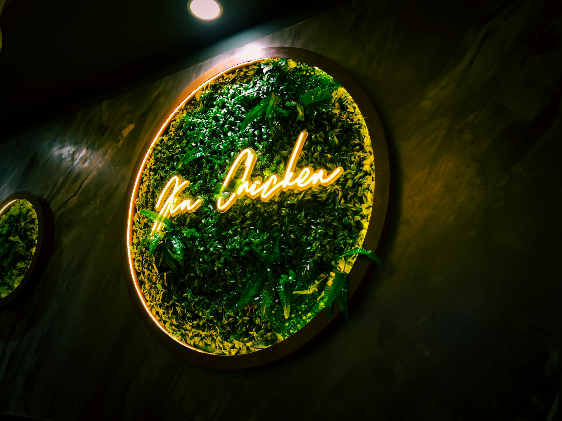 A circular sign with neon lighting displaying the words 'The Chicken' against a lush green plant background. The sign's warm yellow light creates a contrast with the surrounding greenery. The textured wall behind the sign enhances its vibrant appearance.