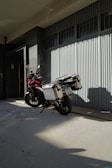 Motorcycle with storage boxes parked on a shaded sidewalk beside a modern building with large windows and a ribbed metal facade.