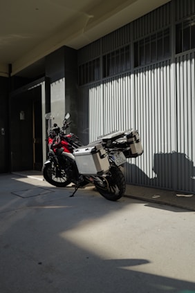 Motorcycle with storage boxes parked on a shaded sidewalk beside a modern building with large windows and a ribbed metal facade.