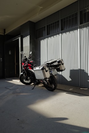 Motorcycle with storage boxes parked on a shaded sidewalk beside a modern building with large windows and a ribbed metal facade.