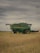 A green agricultural combine harvester is parked in a field of harvested grain under a cloudy sky. The machinery is large, with visible yellow wheels and branding on the side.
