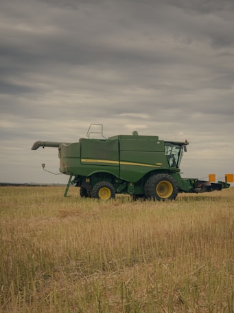 A green agricultural combine harvester is parked in a field of harvested grain under a cloudy sky. The machinery is large, with visible yellow wheels and branding on the side.