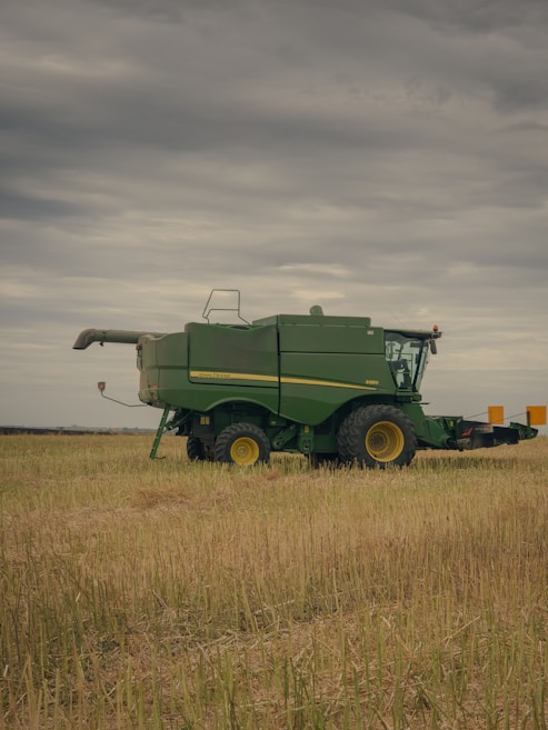 A green agricultural combine harvester is parked in a field of harvested grain under a cloudy sky. The machinery is large, with visible yellow wheels and branding on the side.