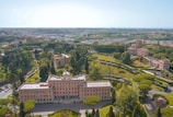 Aerial view of a historical estate surrounded by lush green gardens and trees. The main building is a large, symmetrical structure with a notable architectural style, featuring numerous windows and a central entrance. The landscape includes well-manicured lawns, pathways, and smaller buildings in the surrounding area. The background reveals a sprawling urban cityscape beneath a clear blue sky.