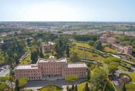 Aerial view of a historical estate surrounded by lush green gardens and trees. The main building is a large, symmetrical structure with a notable architectural style, featuring numerous windows and a central entrance. The landscape includes well-manicured lawns, pathways, and smaller buildings in the surrounding area. The background reveals a sprawling urban cityscape beneath a clear blue sky.