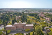 Aerial view of a historical estate surrounded by lush green gardens and trees. The main building is a large, symmetrical structure with a notable architectural style, featuring numerous windows and a central entrance. The landscape includes well-manicured lawns, pathways, and smaller buildings in the surrounding area. The background reveals a sprawling urban cityscape beneath a clear blue sky.