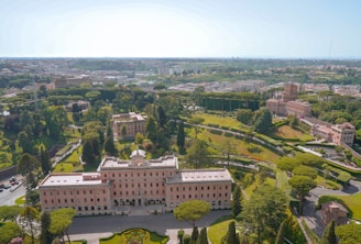 Aerial view of a historical estate surrounded by lush green gardens and trees. The main building is a large, symmetrical structure with a notable architectural style, featuring numerous windows and a central entrance. The landscape includes well-manicured lawns, pathways, and smaller buildings in the surrounding area. The background reveals a sprawling urban cityscape beneath a clear blue sky.