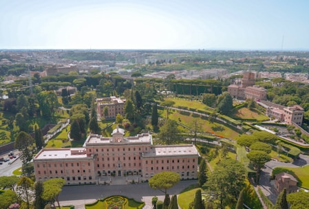 Aerial view of a historical estate surrounded by lush green gardens and trees. The main building is a large, symmetrical structure with a notable architectural style, featuring numerous windows and a central entrance. The landscape includes well-manicured lawns, pathways, and smaller buildings in the surrounding area. The background reveals a sprawling urban cityscape beneath a clear blue sky.