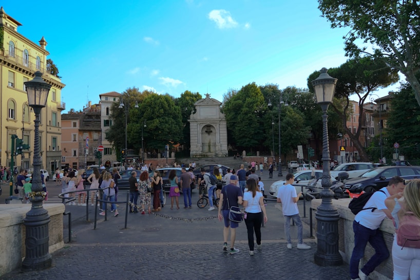 The scene captures a bustling city square with people walking and socializing near a historic monument surrounded by lush trees. The area is vibrant with pedestrians, cars, and scooters passing by, while antique street lamps add a classic touch. The architecture of surrounding buildings showcases a blend of historical and residential styles.