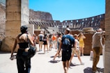 Tourists walk through the ancient ruins of a large amphitheater, with stone walls and arches visible in the background. The scene is bustling with people capturing photos and exploring the historical architecture. The sky is clear and bright, adding to the vibrancy of the setting.