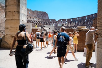 Tourists walk through the ancient ruins of a large amphitheater, with stone walls and arches visible in the background. The scene is bustling with people capturing photos and exploring the historical architecture. The sky is clear and bright, adding to the vibrancy of the setting.