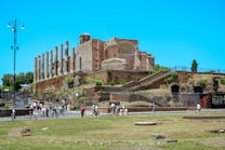 Ancient Roman ruins with a series of tall stone columns extending alongside aged brick structures. People are walking along a pathway at the base of the ruins. The sky is clear and bright, enhancing the historic urban landscape.