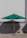 A large green outdoor umbrella stands on a paved surface, casting a shadow against a patterned stone or tiled wall. Two metal chairs are positioned on either side of the umbrella base.