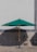 A large green outdoor umbrella stands on a paved surface, casting a shadow against a patterned stone or tiled wall. Two metal chairs are positioned on either side of the umbrella base.
