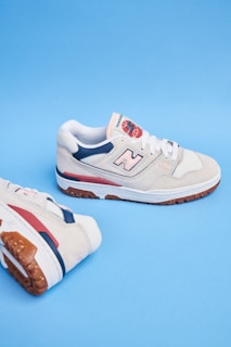 A pair of beige athletic shoes with white laces and blue and red accents. The shoes feature a noticeable logo on the side and are set against a solid blue background.