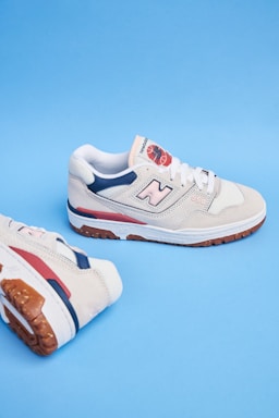 A pair of beige athletic shoes with white laces and blue and red accents. The shoes feature a noticeable logo on the side and are set against a solid blue background.