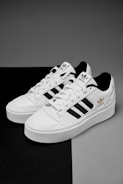 A pair of white sneakers with black stripes and gold branding details are placed on a contrasting black and gray surface. The shoes feature a classic design with white laces and branding elements on the tongue and side.