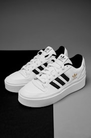 A pair of white sneakers with black stripes and gold branding details are placed on a contrasting black and gray surface. The shoes feature a classic design with white laces and branding elements on the tongue and side.