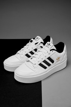 A pair of white sneakers with black stripes and gold branding details are placed on a contrasting black and gray surface. The shoes feature a classic design with white laces and branding elements on the tongue and side.
