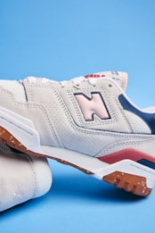 A close-up of a stylish sneaker with a beige and pastel pink color scheme. The shoe features perforated suede material, a prominent logo on the side, and contrasting details in navy blue and red. The sole has a rugged texture in a gum color.