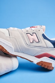 A close-up of a stylish sneaker with a beige and pastel pink color scheme. The shoe features perforated suede material, a prominent logo on the side, and contrasting details in navy blue and red. The sole has a rugged texture in a gum color.